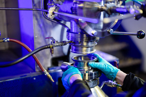 Close-up of gloved hands adjusting a fitting on the pilot reactor, lit in purple