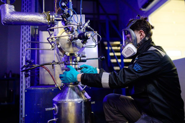 A technician in a respirator mask adjusts a valve on a stainless steel pilot reactor